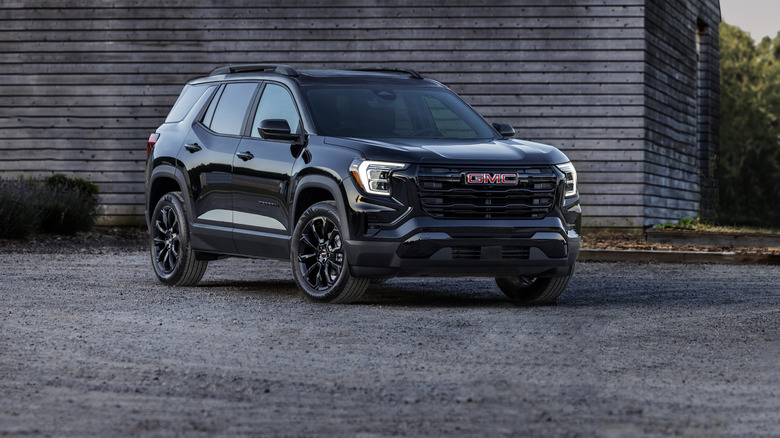 Black 2025 GMC Terrain Elevation SUV seen from front right against a backdrop of a wooden structure