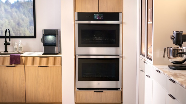 A GE stainless steel double wall oven in a modern kitchen, with a small touchscreen display at the top and a countertop ice maker and stand mixer visible on either side.