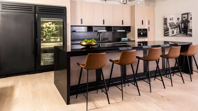 A black True Refrigeration column unit with a glass door sitting against light wood cabinetry in a modern kitchen.
