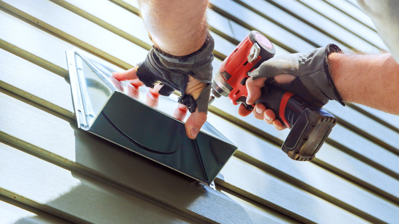 A man uses a cordless drill to install a vent cover on the side of a home.