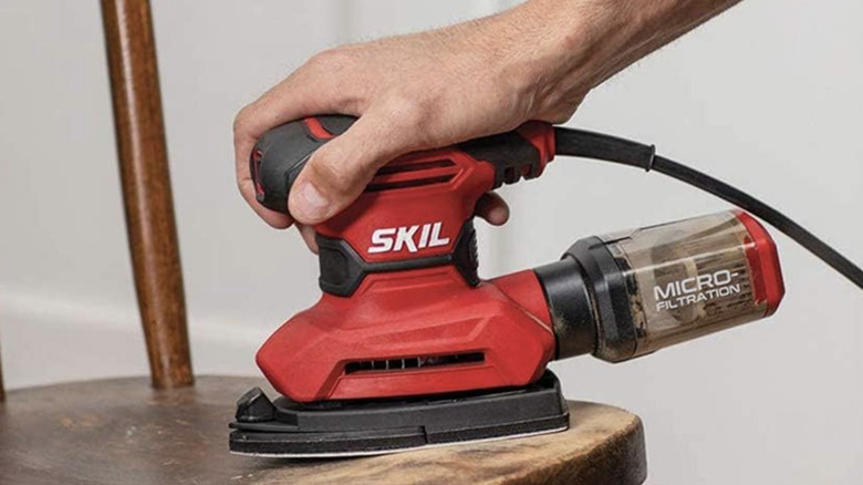 A man is shown sanding a SKIL Corded Multi-Function Detail Sander to sand a wooden chair.
