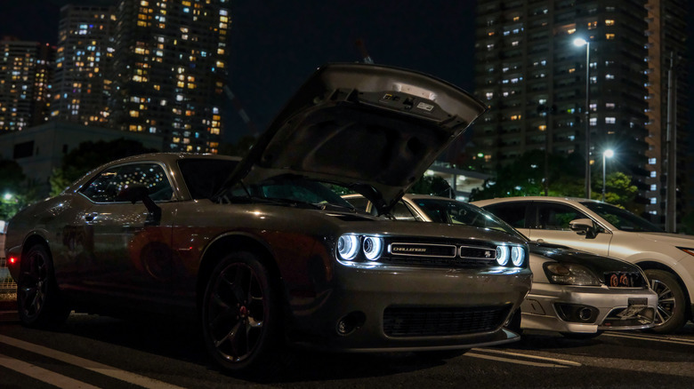 A silver Dodge Challenger, headlights illuminated, with its hood open at night