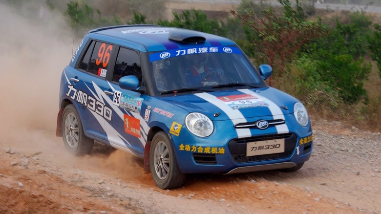 A blue Lifan 330 car going rally racing on a dirt road, covered with sponsor logos