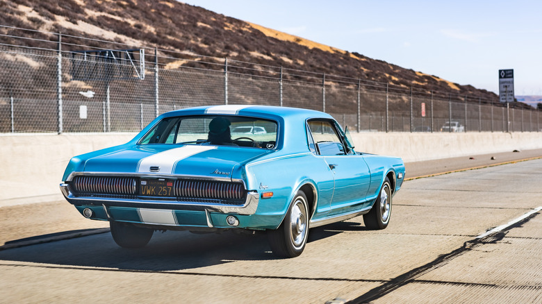 a 1967 Mercury Cougar in blue with white racing stripes, driving along a highway beside a chain-link fence