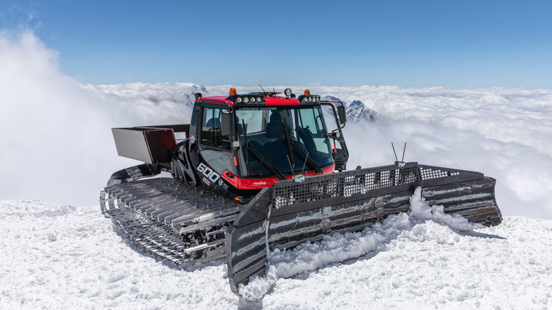 A snowcat clearing snow from a path with mountains and clouds in the rearview.