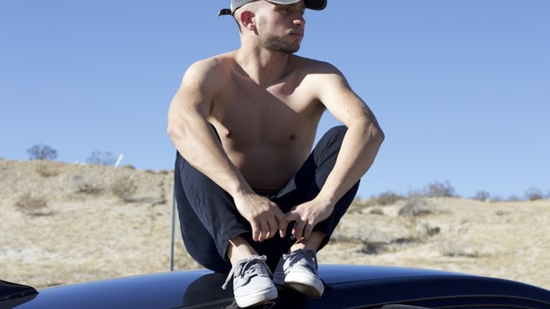 topless man in blue denim jeans and brown cowboy hat sitting on black car during daytime.