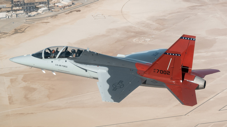 T-7A Red Hawk jet seen from above left flying above a desert airbase
