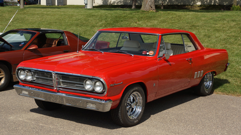 Red 1965 Acadian Beaumont coupe seen from left front parked at a car show next to 1980s Camaro