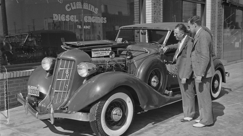 Fred Duesenberg examines a diesel-powered automobile with its hood open, showcasing Clessie Cummins's pioneering diesel engine.
