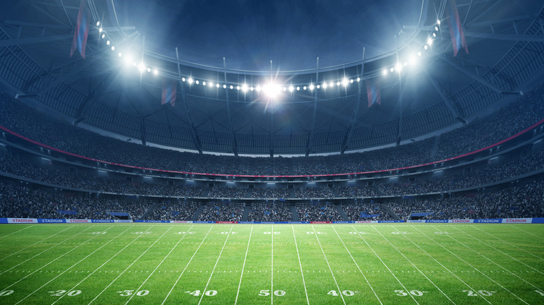A brightly lit football stadium filled with fans, showing a green field marked with yard lines and the 50-yard line at the center.