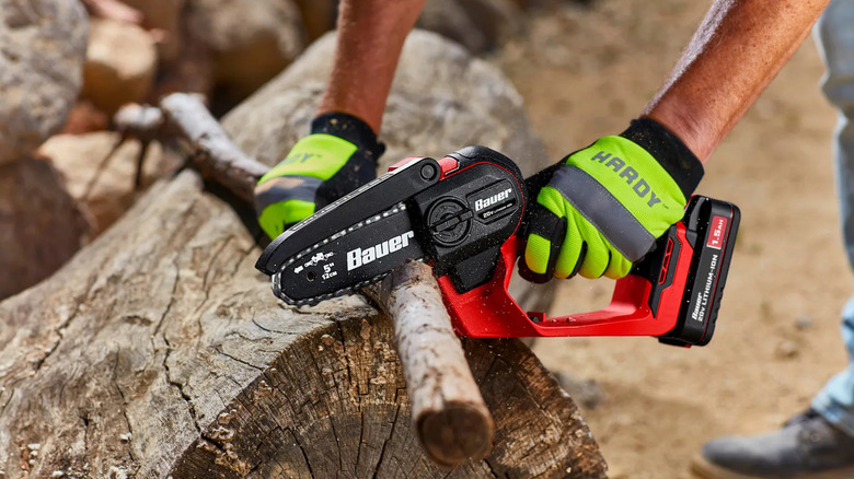 A person using a Bauer 20V cordless mini chainsaw to cut a small branch resting on a large tree stump.