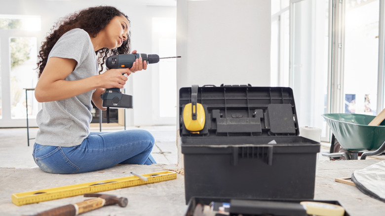 A woman changing a drill bit while sitting on a step, with a tool box and some hand tools in the foreground