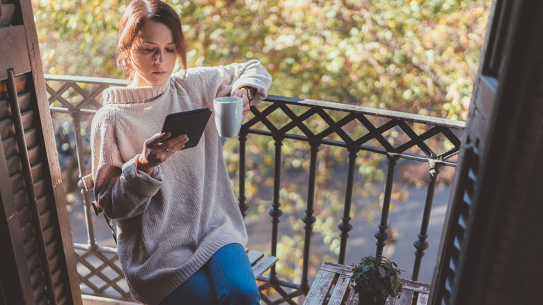 A person drinking coffee and looking at an e-reader on a cozy balcony