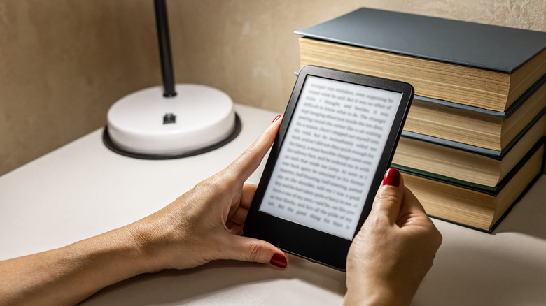 A person holding an e-reader in front of some books on a table, next to a lamp