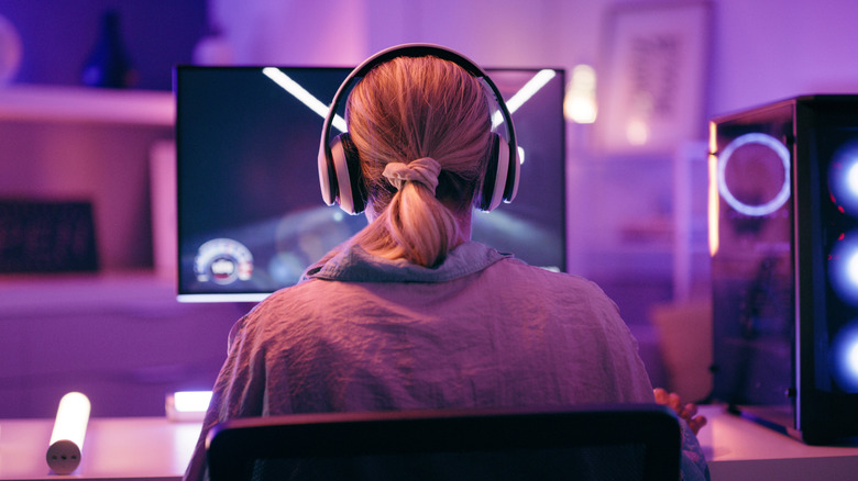 A person wearing a pair of headphones while sitting on a desk with a monitor and computer case next to it, bathed in neon light
