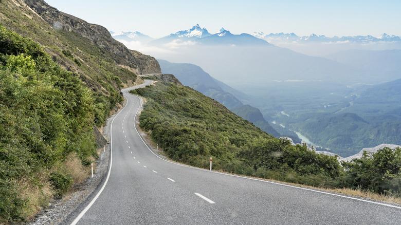 Winding mountain road in Washington State, curving along a steep hillside with lush greenery, overlooking a misty valley and snow-capped peaks in the distance.
