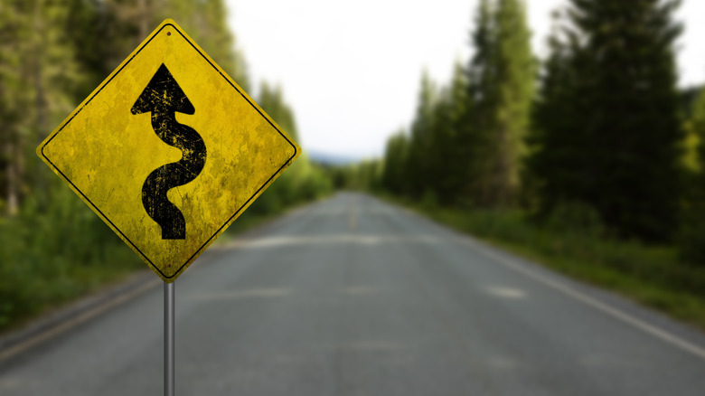 Close-up of a weathered winding road warning sign on a quiet, tree-lined highway.