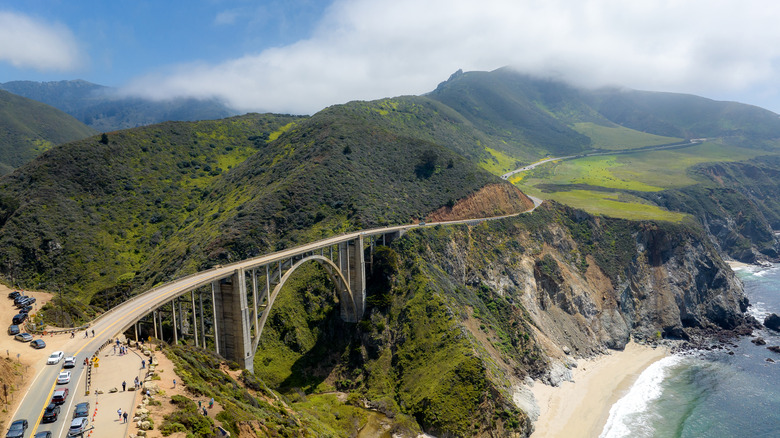 Aerial view of Bixby Creek Bridge along California's Pacific Coast Highway, with a sandy beach below and misty mountains in the distance