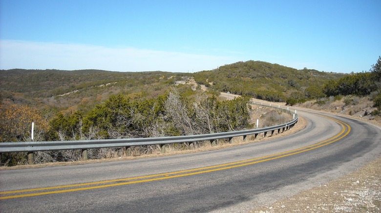 Curving two-lane road through the Texas Hill Country, lined with guardrails and surrounded by rolling hills, dry brush, and scattered trees under a clear blue sky.