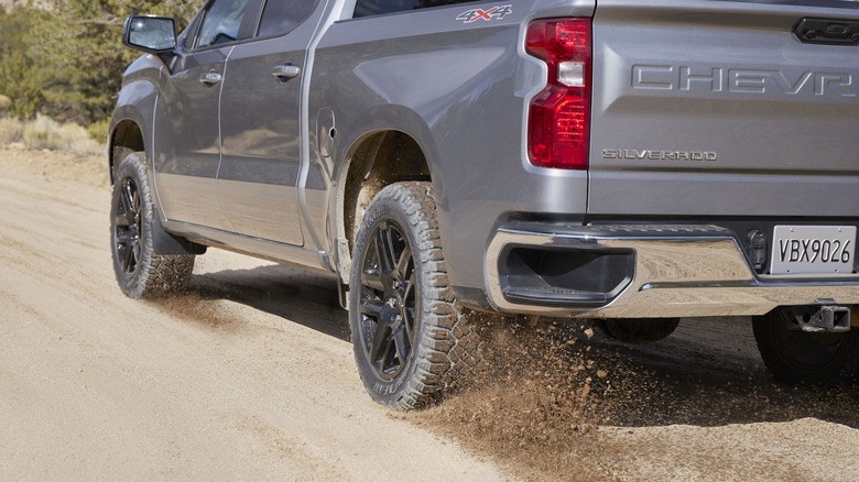The Goodyear Wrangler DuraTrac RT all-terrain tires attached to a pickup truck that's driving down a dirt road, with the tires throwing dirt behind them