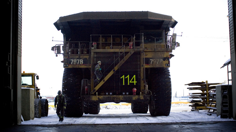 A massive CAT 797b mining truck parked facing the camera in a large industrial garage, with two workers in safety gear nearby.