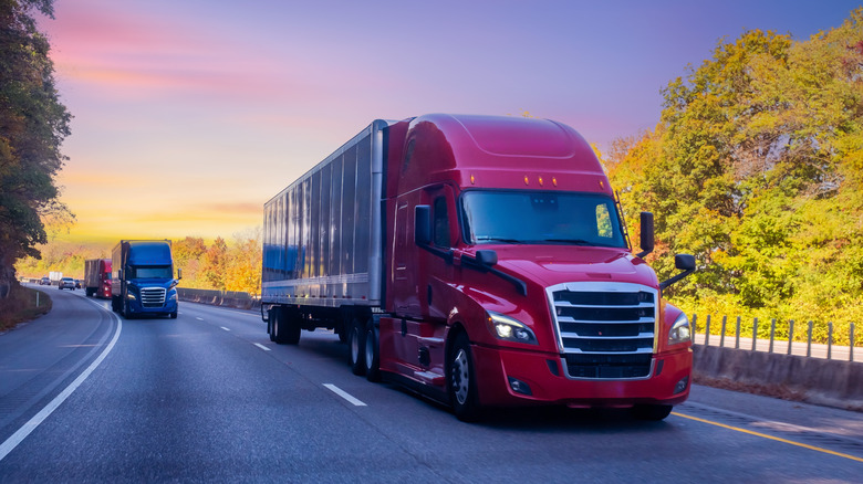 A line of semi-trucks driving on a highway during sunset, with colorful skies and autumn trees in the background.