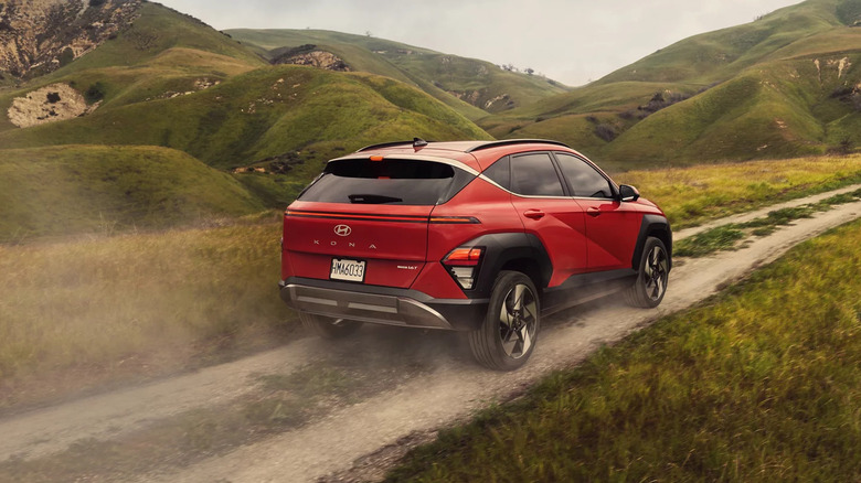 2025 Hyundai Kona in red driving on a dirt trail through rolling green hills, kicking up dust as it navigates a scenic countryside landscape.