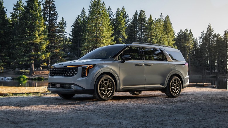 A grey metallic Kia Carnival that's standing still against the backdrop of a forest