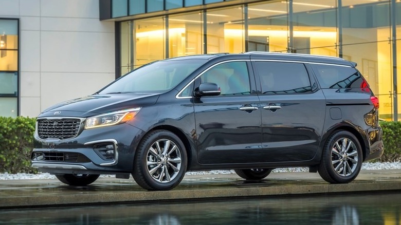 A metallic grey Kia Sedona 2021 against the backdrop of a glass building, parked horizontally near a water feature