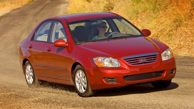 A blue 2007 Kia Spectra against a forested background, parked diagonally on a road.