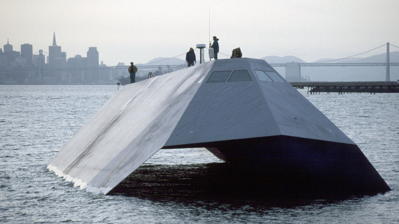 Lockheed Sea Shadow stealth ship seen from in front in a city harbor with buildings and bridge in background