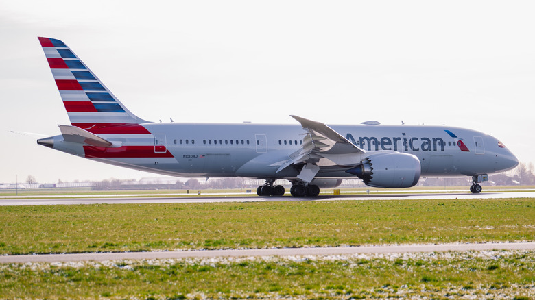A Boeing 787-8 airliner from the fleet of American Airlines, taxiing down a runway, left to right, airport buildings visible in the background.