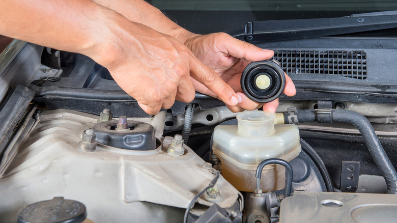 Person checking the brake fluid reservoir under a car's hood, while pointing to the fluid inside.