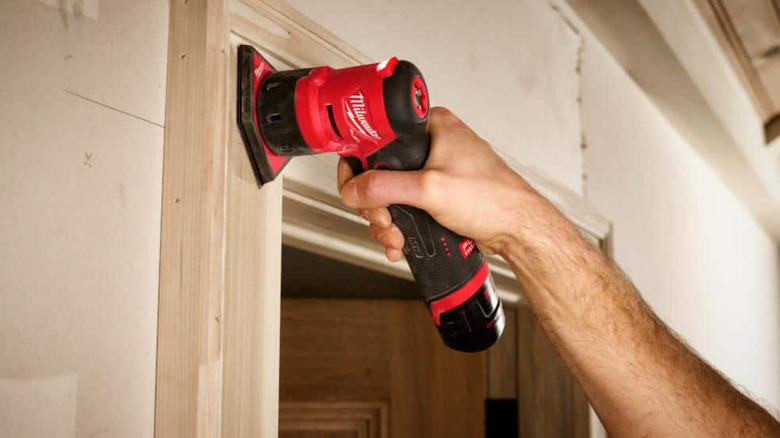 A person using a Milwaukee M12 Fuel Orbital Detail Sander on a door frame.