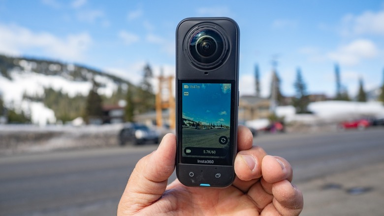 The Insta360 X5 held in a hand with a view of a mountain road shown in the background and on the display.