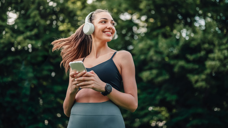 A woman in a park with headphones on, holding a smartphone.