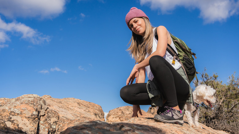A hiker crouching on a rock with dog and light pack on her back.