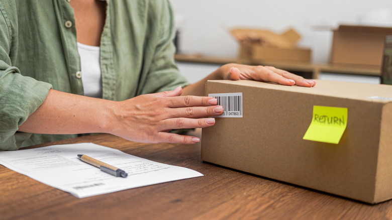 A woman sitting at a wooden table, placing her hands on a brown cardboard box with a yellow sticker on the side.