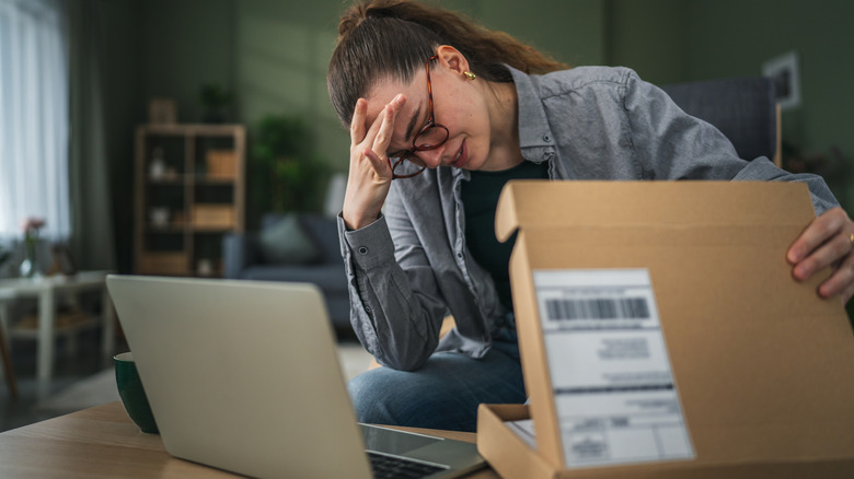 A woman with a disappointed look holding an open brown parcel with a laptop open in front of her.