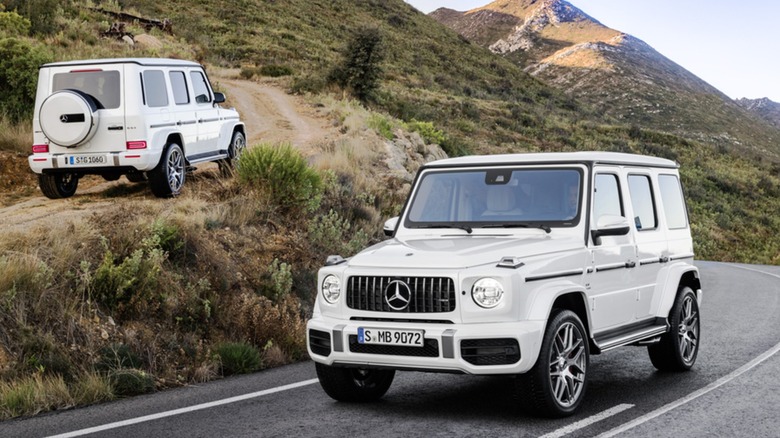Two white 2019 Mercedes AMG G63s; One is sitting on the road facing the camera, and the other is driving up a dirt road in the opposite direction.