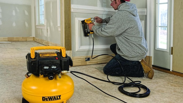 A man uses a compressed air stapler on a wall. A DeWalt air compressor can be seen nearby.