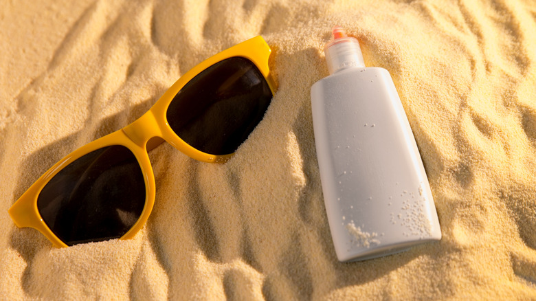 An unlabeled sunscreen bottle and yellow sunglasses on sand.