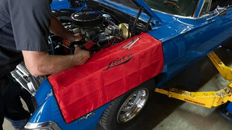 A mechanic working on the engine of a classic blue muscle car sitting low on a lift, with a red Lisle fender cover prominently displayed.