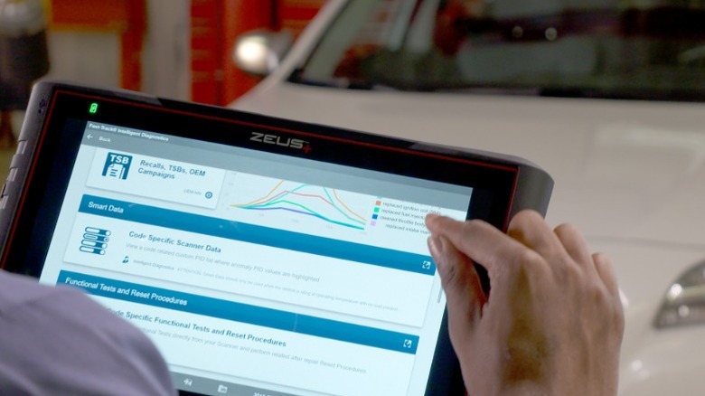 A close-up of the Snap-on Zeus automotive scanner with a hand tapping on the screen and a white car in the background.
