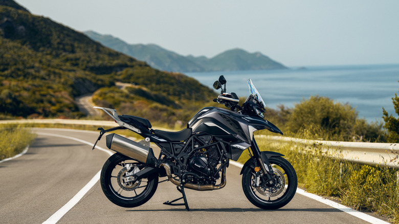 A Benelli TRK 702 adventure motorcycle parked on a scenic winding mountain road with the sea and green hills in the background, under clear skies.