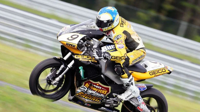 A rider in yellow and black racing gear leans into a corner on a Buell race motorcycle with number 9, heavily branded with GEICO Powersports and other sponsor logos, on a racetrack.