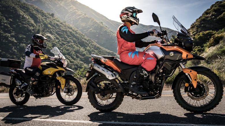 Two bikers riding CSC motorcycles down a mountain path, dropoff visible in the background, right side view