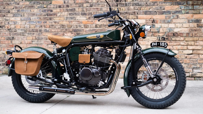 A vintage-styled green Janus Halcyon 450 motorcycle with tan leather seat and saddlebags, parked on a sidewalk against a rustic brick wall.