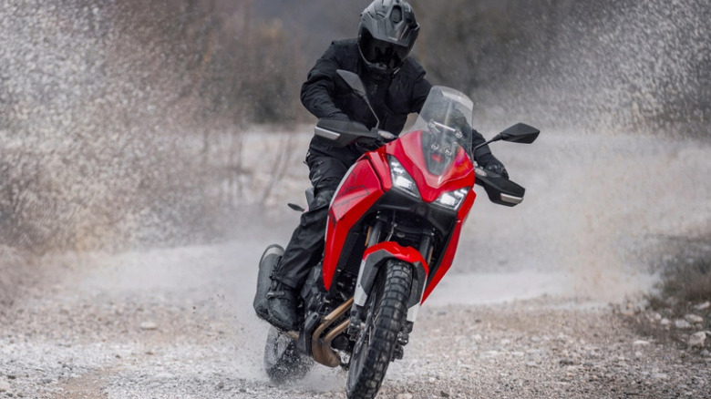 A rider in black adventure gear powers a red Moto Morini adventure motorcycle through a gravel trail, kicking up rocks and dust while navigating a turn in an off-road setting.
