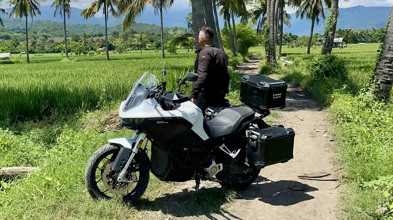 A rider on a white Zero DSR/X electric adventure motorcycle is parked on a dirt path surrounded by lush green rice fields and tall palm trees, with mountains in the background under a bright blue sky.
